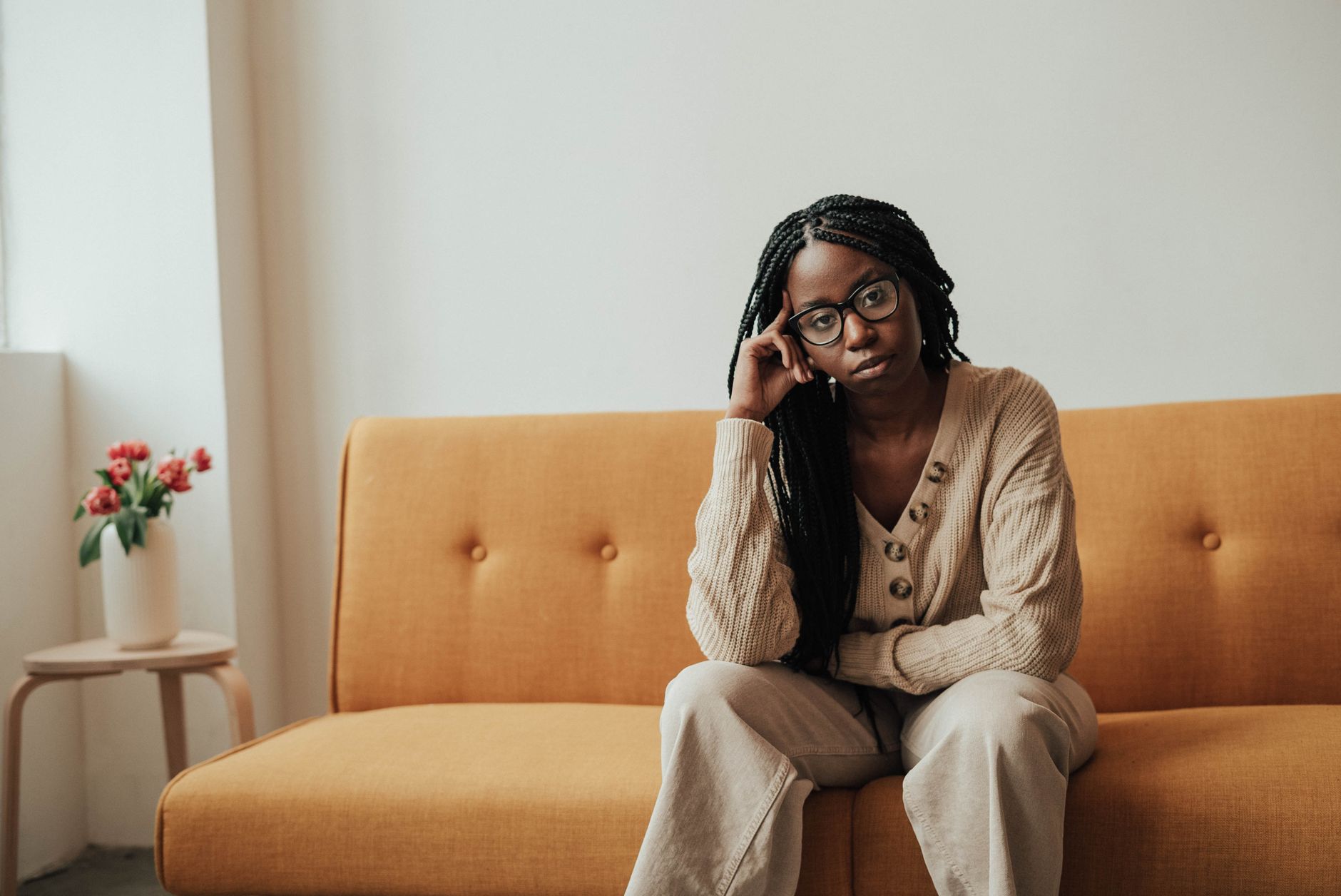 A woman with braids and glasses sits contemplatively on a mustard couch