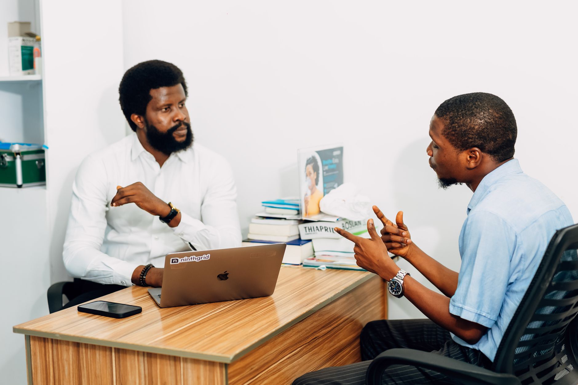Two men discuss event planning at a desk with a laptop and books