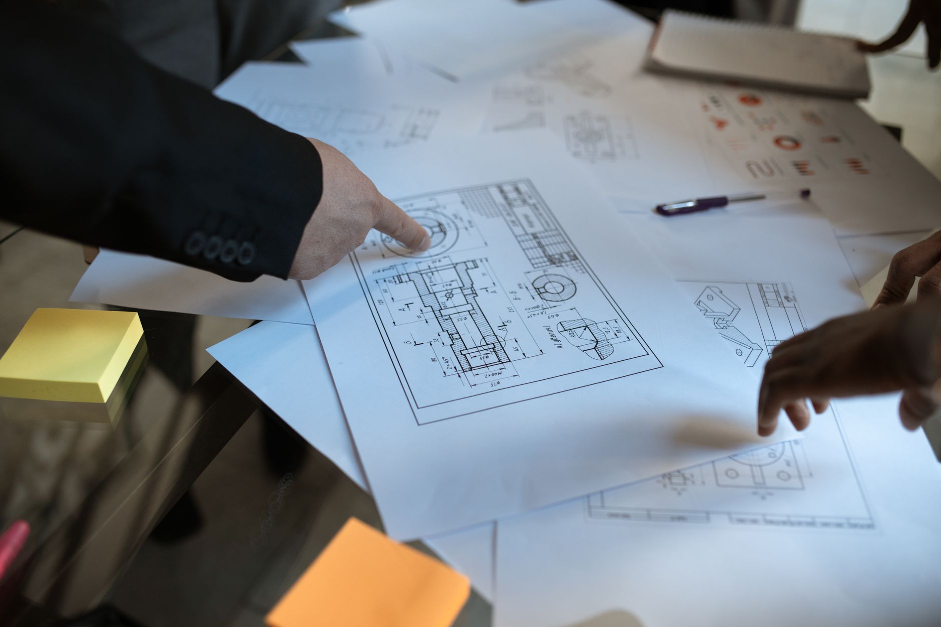 A person's hand points at engineering blueprints on a table with other papers