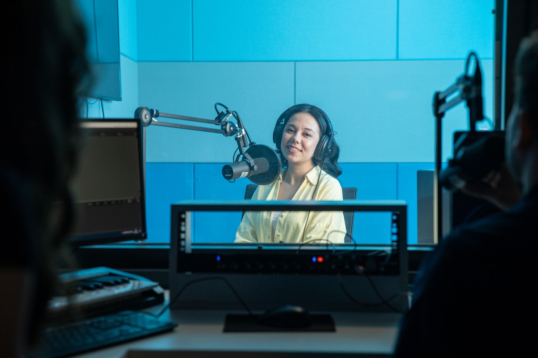 A woman wearing headphones smiles behind a microphone inside a blue recording studio