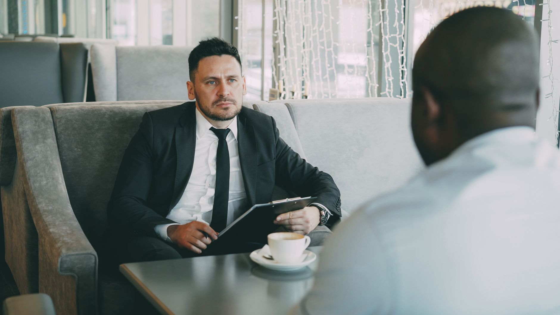 A man in a suit holds a clipboard, negotiating with another man in a cafe