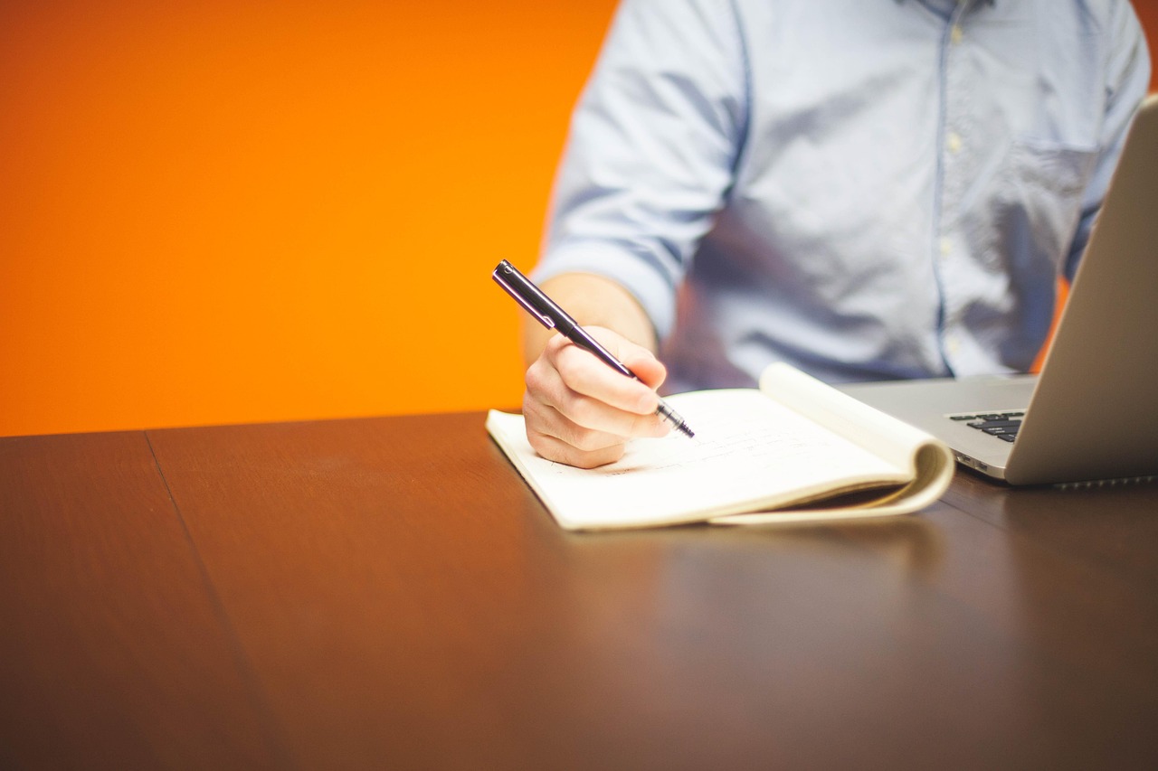A person writes notes in a notebook next to a laptop on a desk