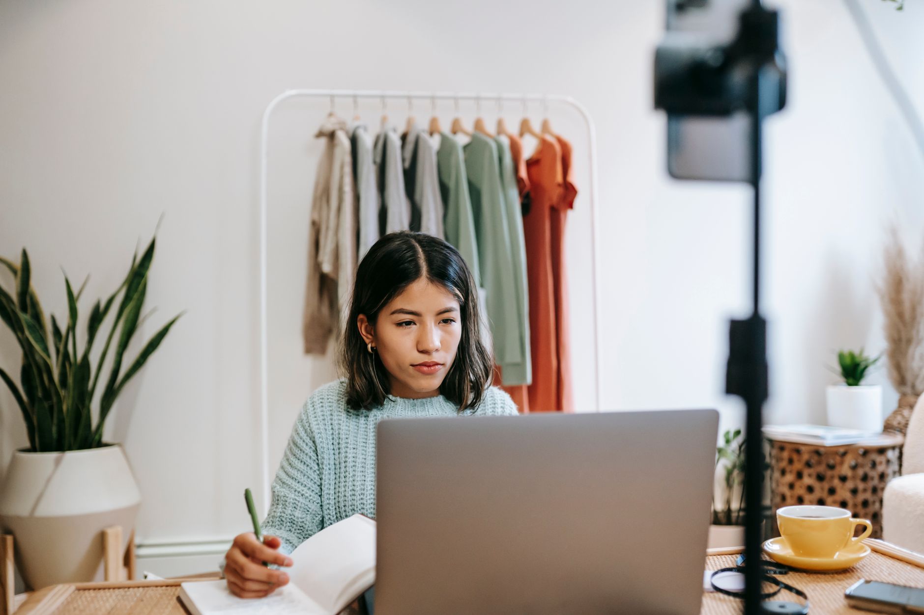 A woman holding a pen and notebook looks at an open laptop