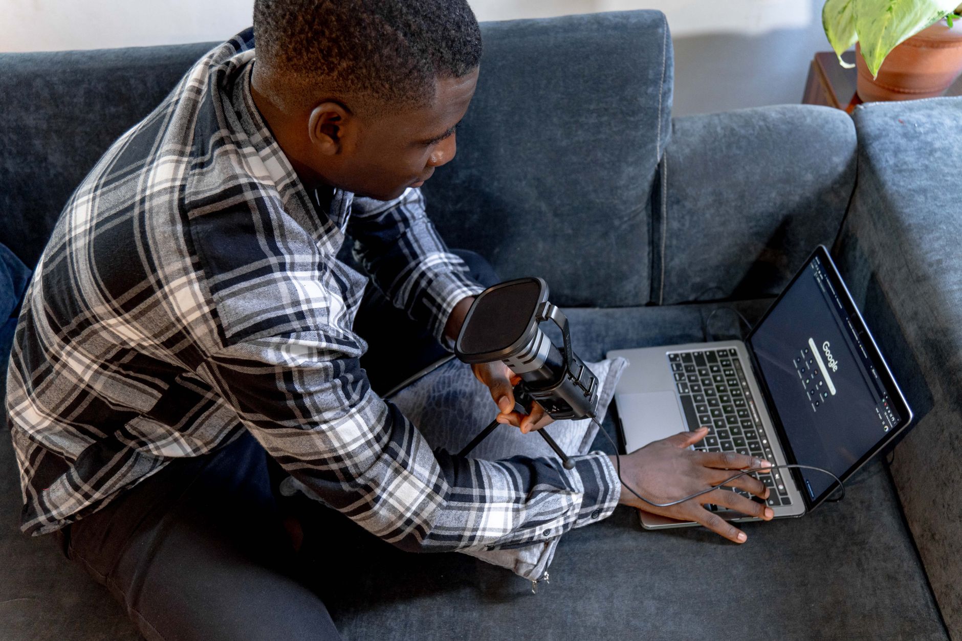 A man on a couch holds a microphone while using a laptop
