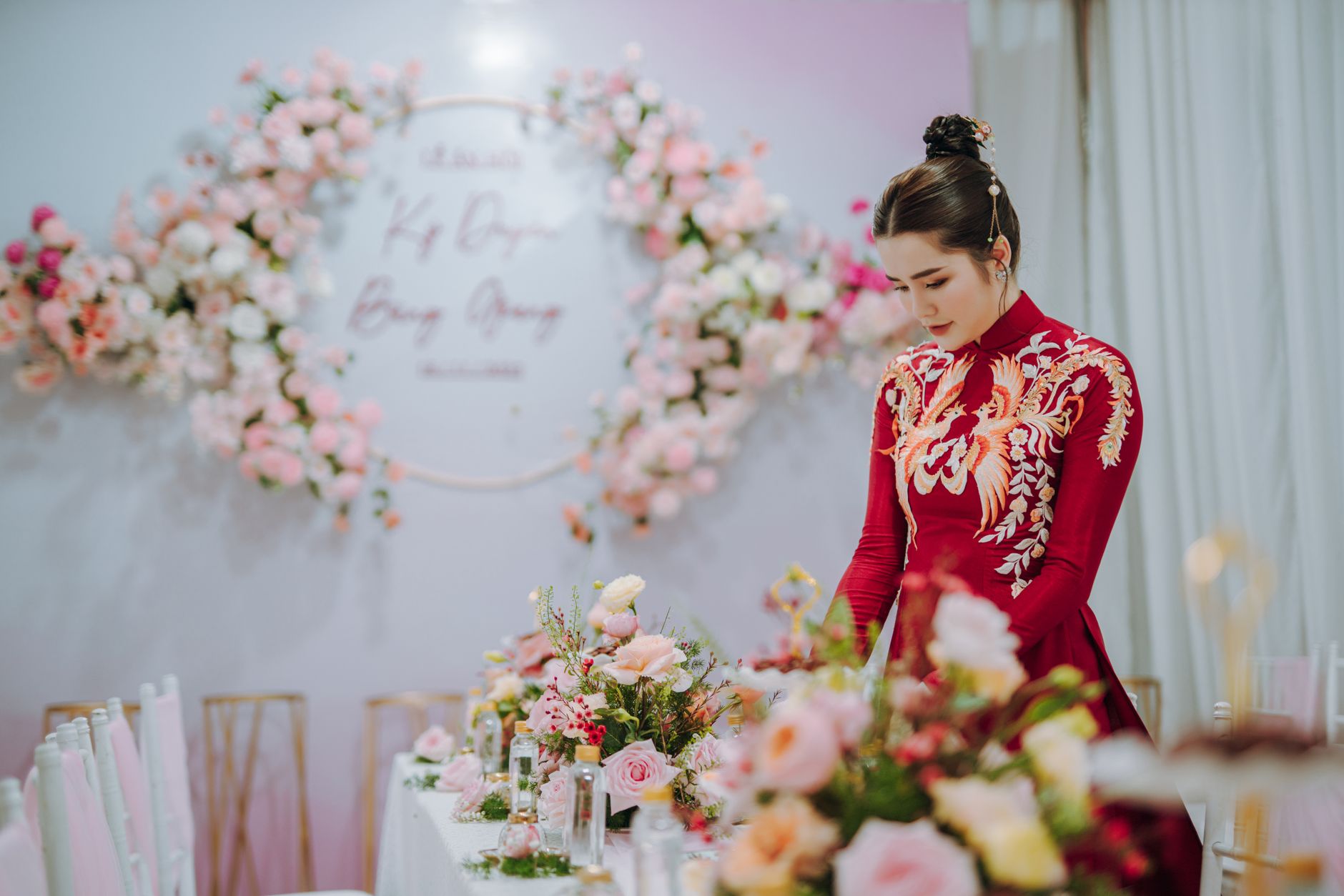 Woman in embroidered red dress stands by a table adorned with pink flowers