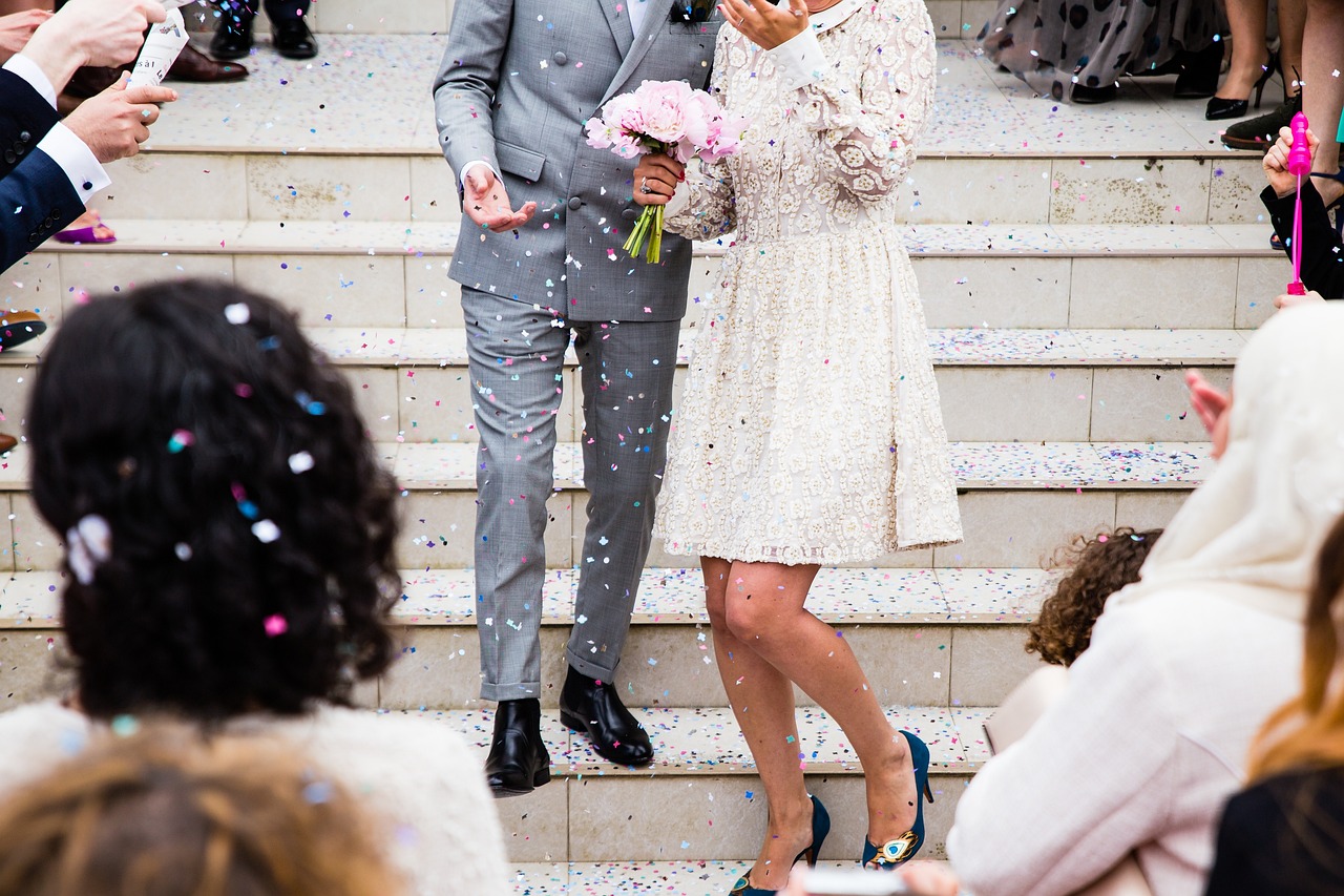 A couple stands on stairs, showered in colorful confetti with a pink bouquet