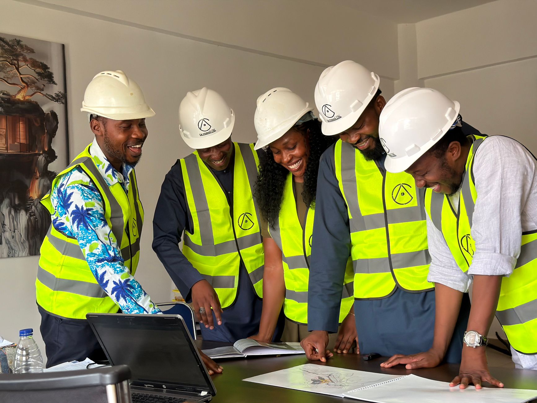Five smiling people in hard hats and safety vests review documents and a laptop