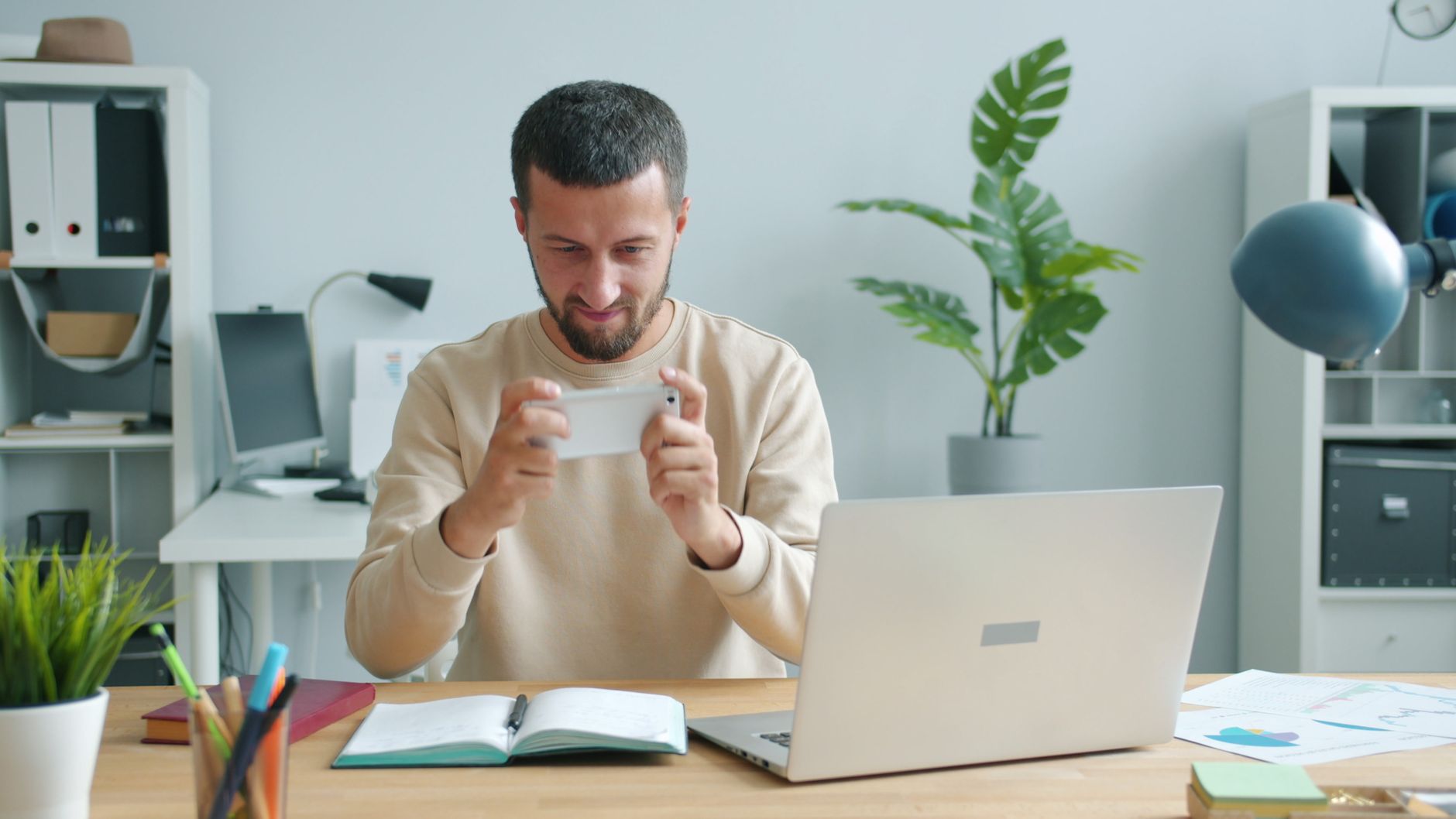 A bearded man sits at a wooden desk holding a smartphone horizontally