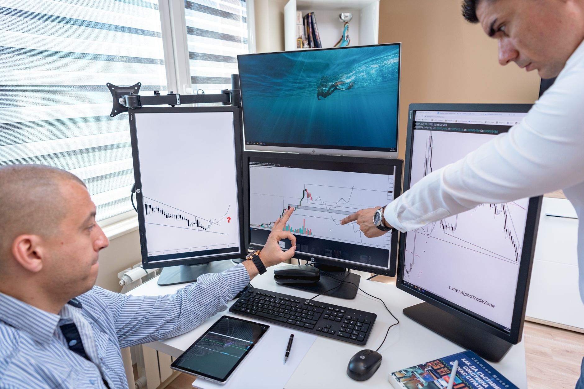 Two men in an office point at computer monitors showing financial charts