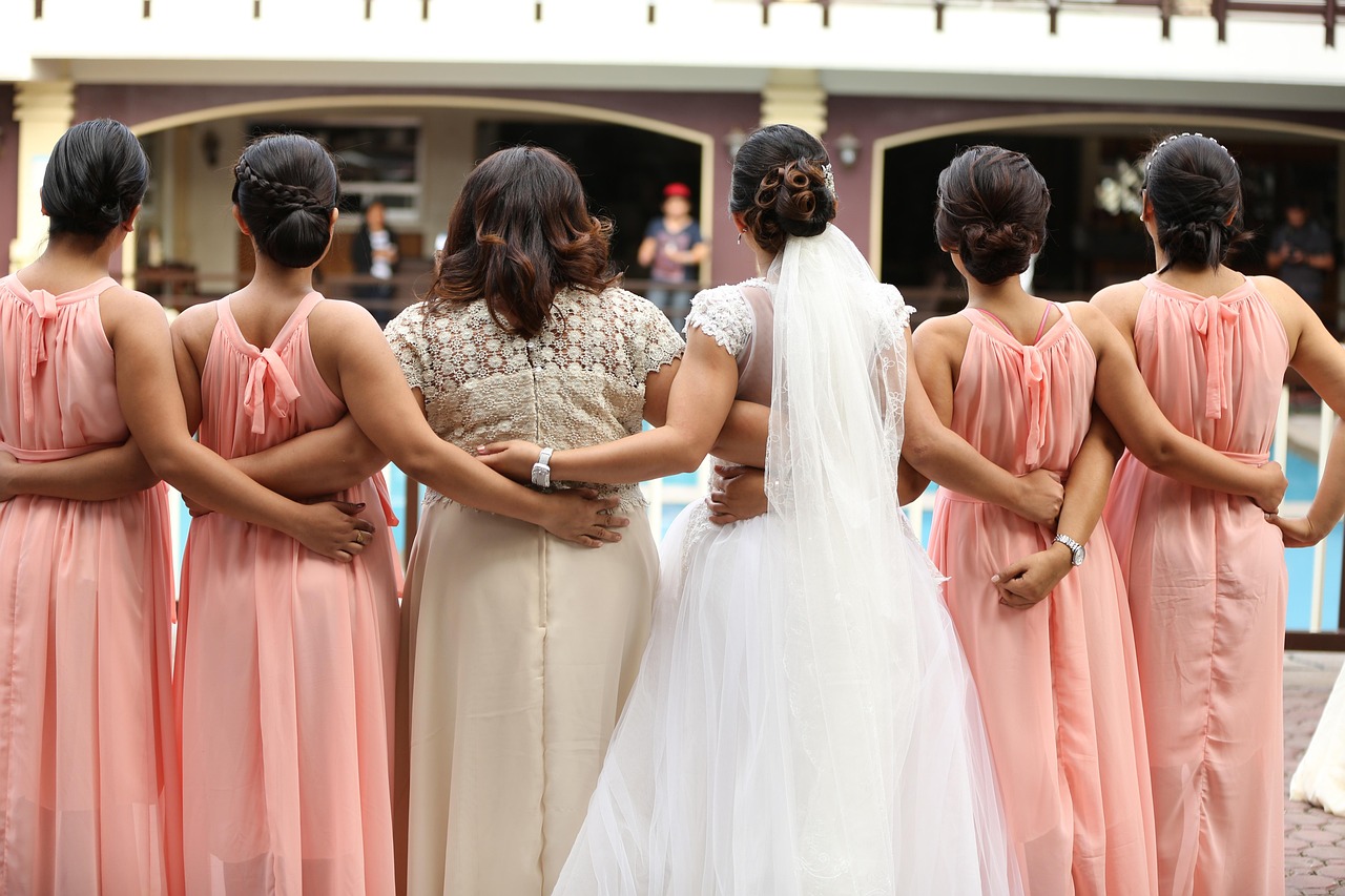 Bride and bridesmaids in peach and cream dresses, arms linked, from behind