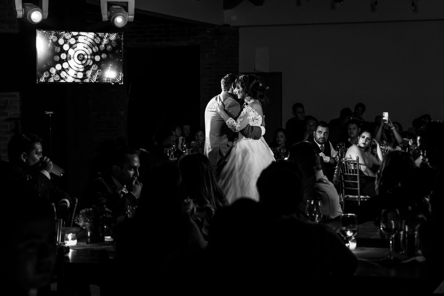 A bride and groom share a first dance under spotlights, guests watching