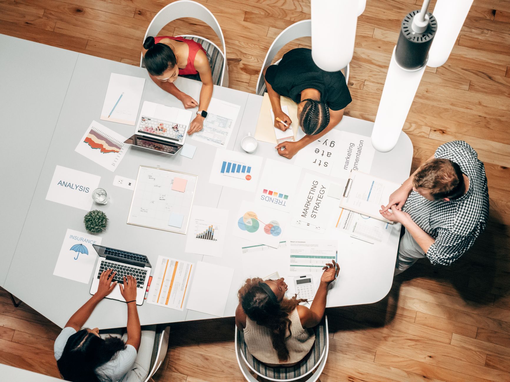 Five people sit and stand around a table covered with papers and laptops