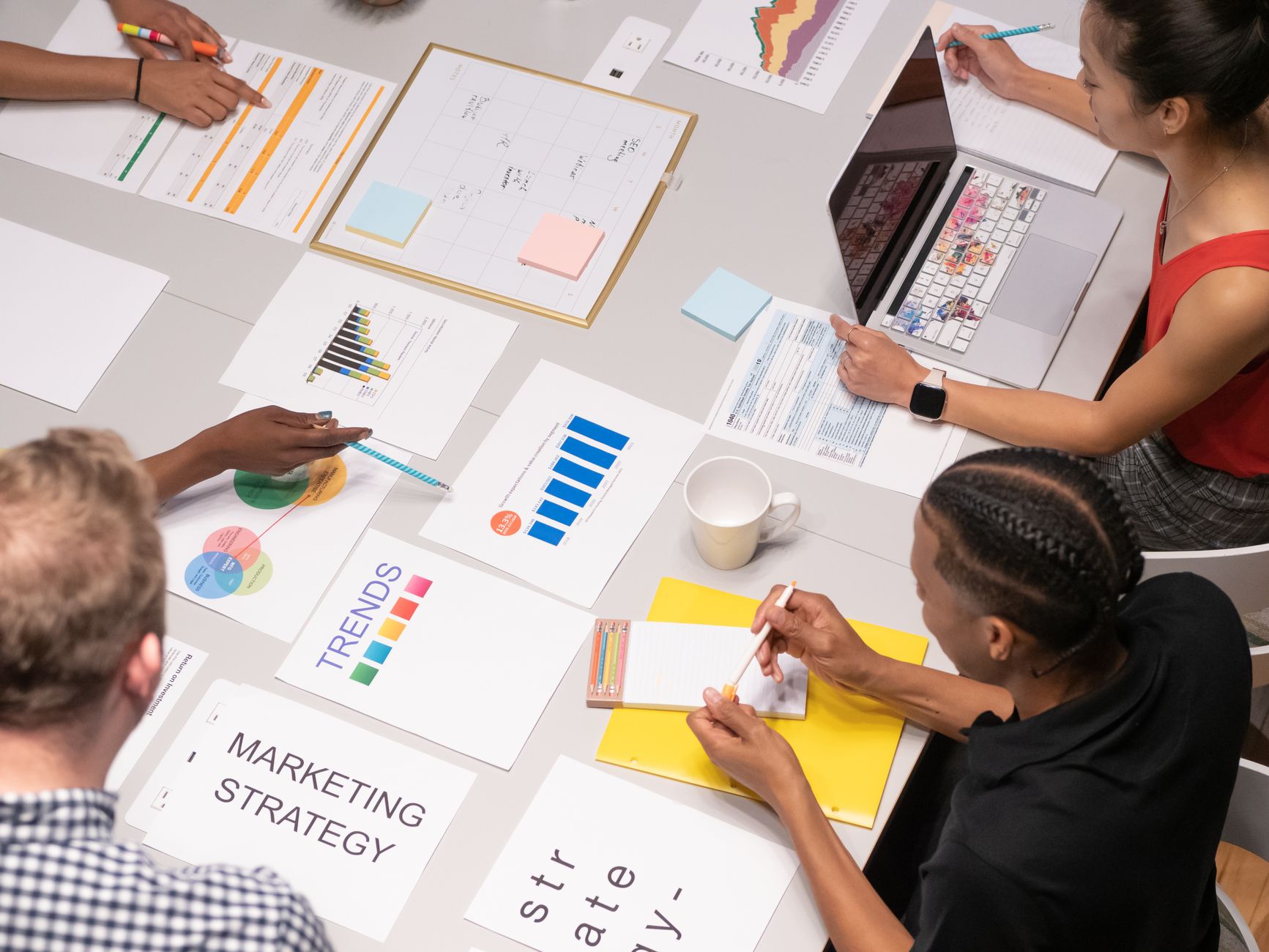 People collaborate at a table covered with marketing documents, charts, and a laptop