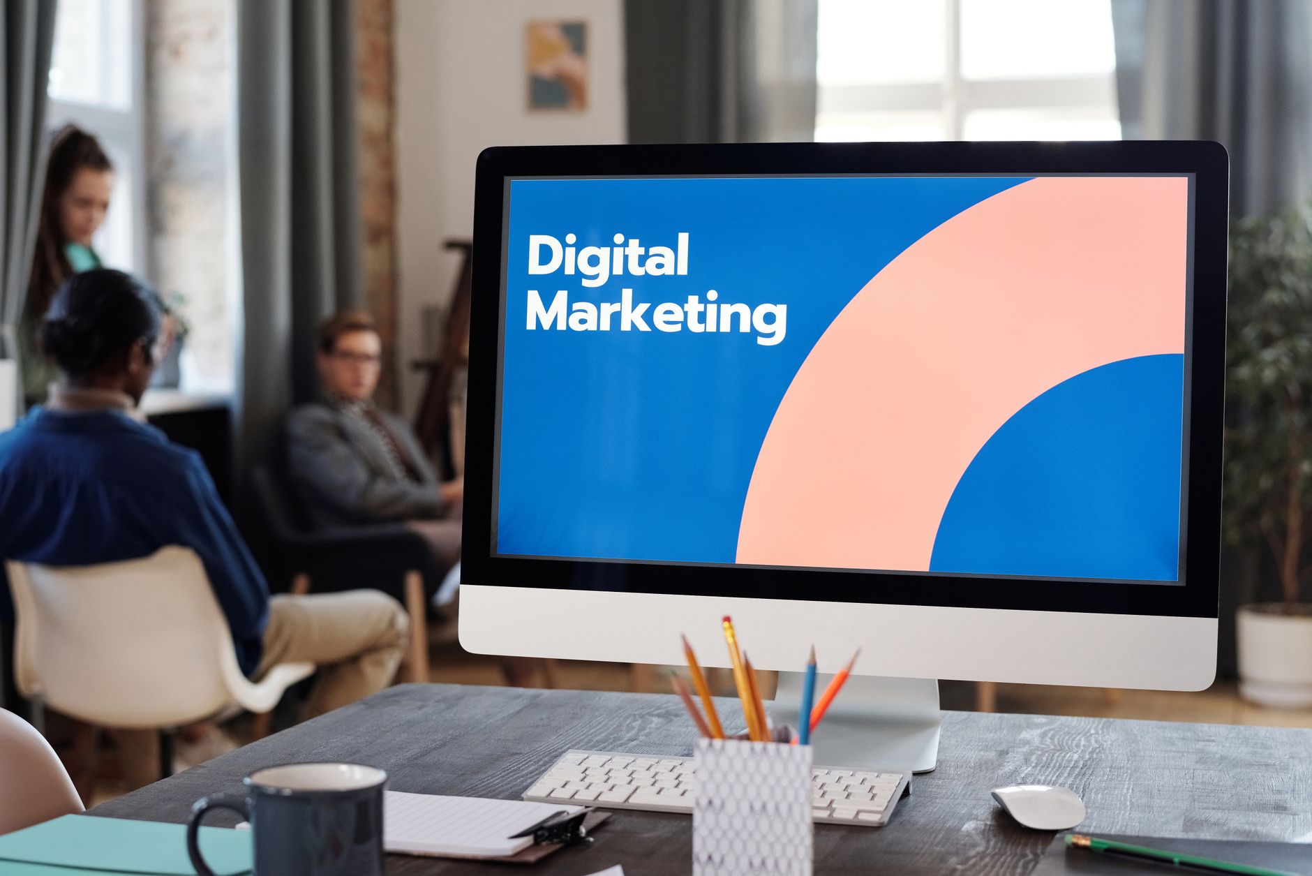 Computer screen displays "Digital Marketing" text on a desk with blurred people behind it