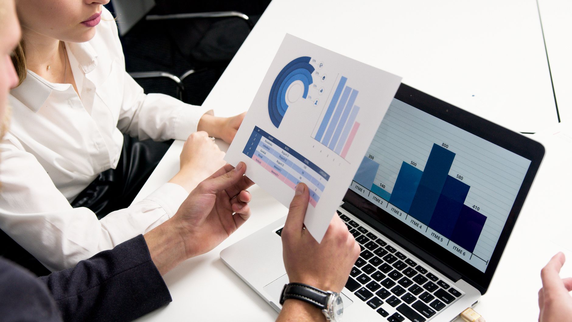 Two professionals analyze data from a printed chart and a laptop displaying a bar graph