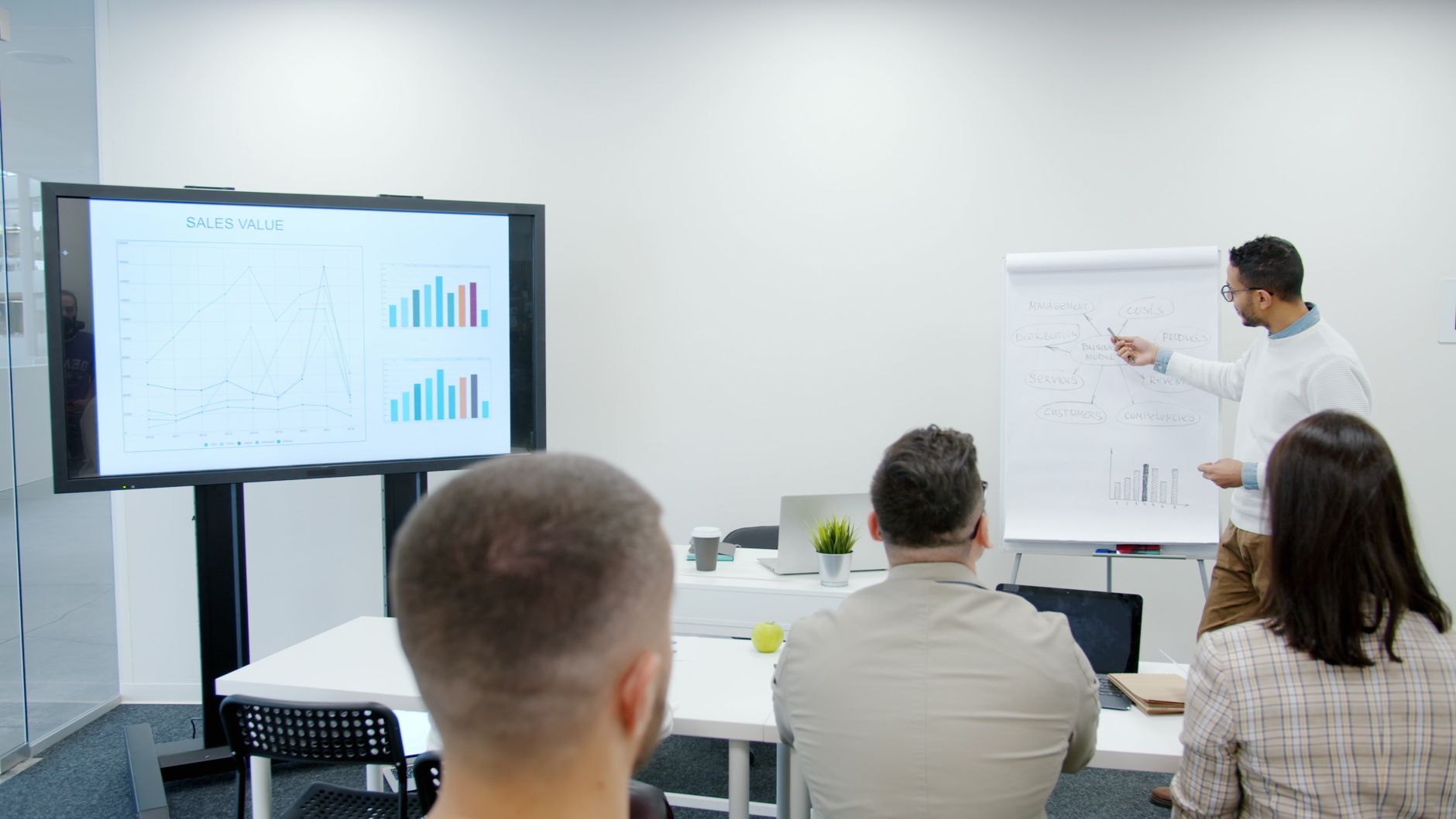 A man presents to three seated people beside a monitor showing charts
