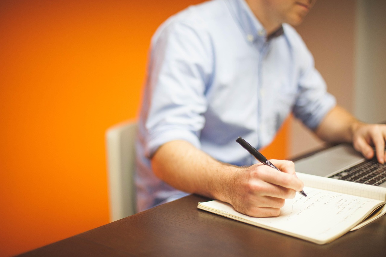 A person writes notes in a notebook while working on a laptop at a desk