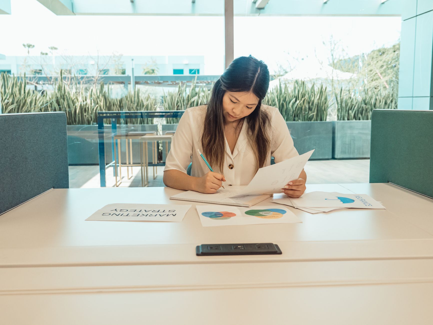 A woman sits at a desk reviewing printed papers with a pencil