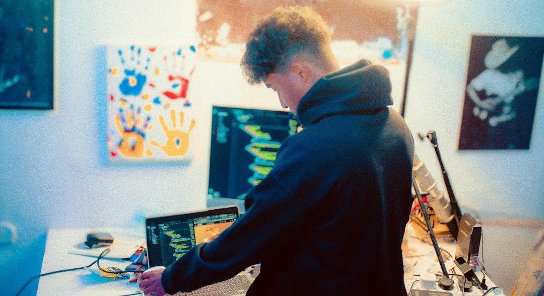 A man in a hoodie stands at a desk looking at computer screens