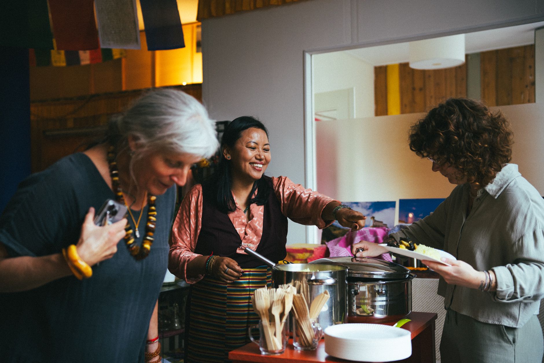 Three women smiling and serving food together from large pots during an event