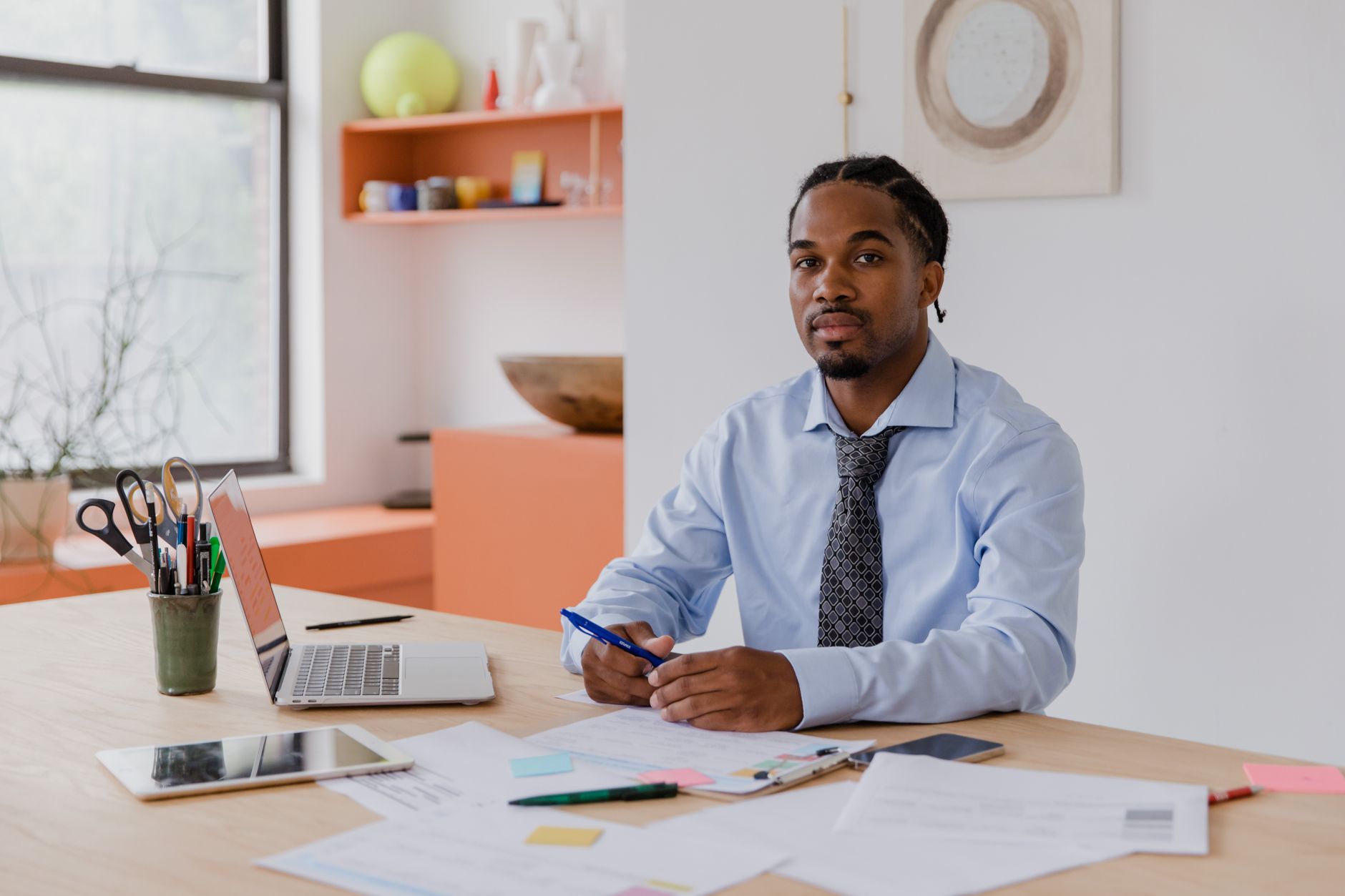 A man in a shirt and tie reviewing documents on a desk with a laptop