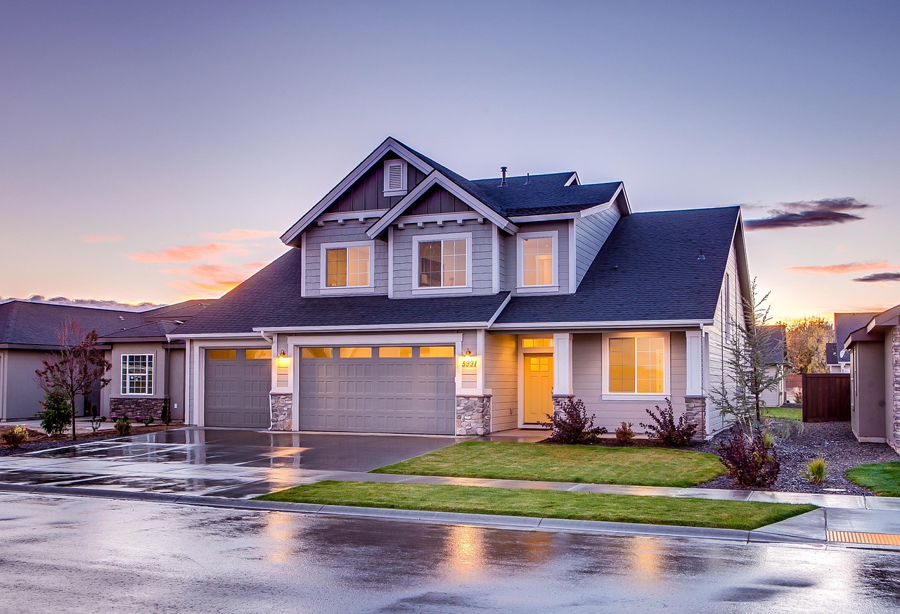 A large, modern suburban house at dusk with glowing interior lights reflected on a wet street