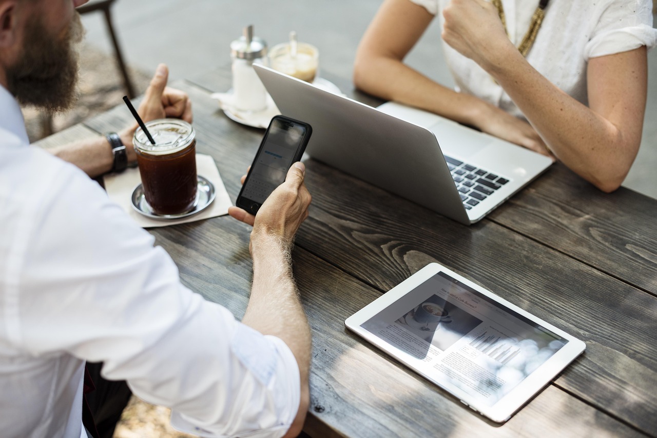 Two people consult mobile devices, laptop, and tablet on a wooden table