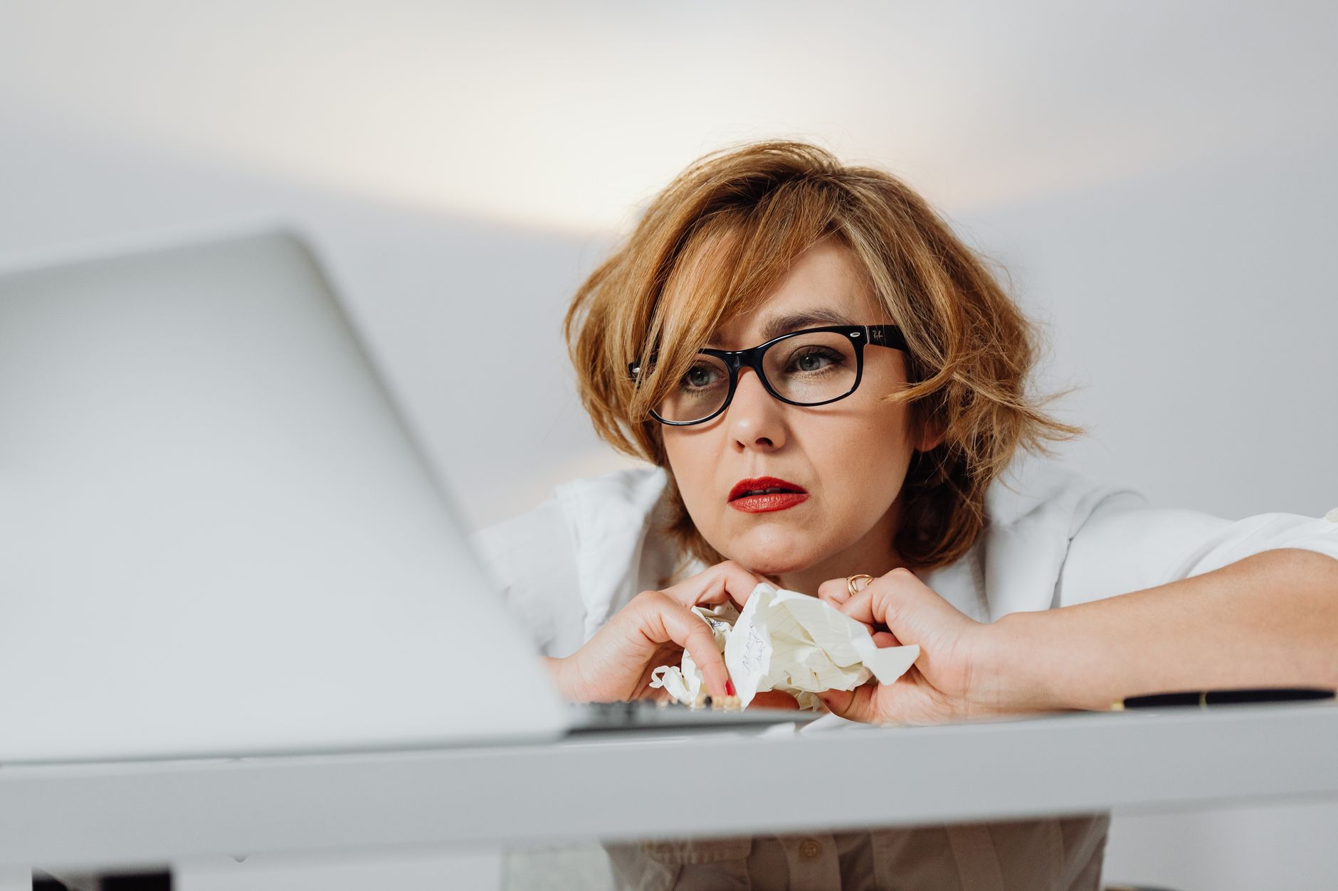 A woman wearing glasses holds crumpled paper while looking at a laptop screen