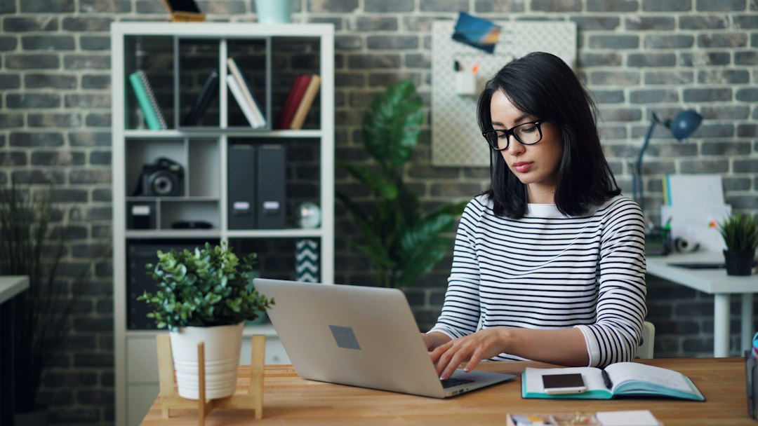A woman wearing glasses types on a laptop at a wooden desk