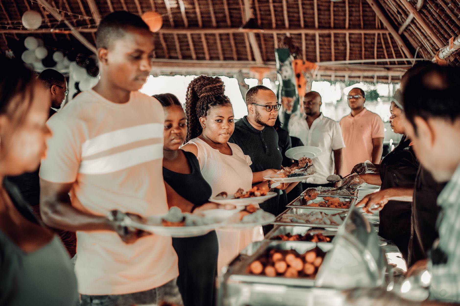 People fill plates with African food from a buffet line under a thatched roof