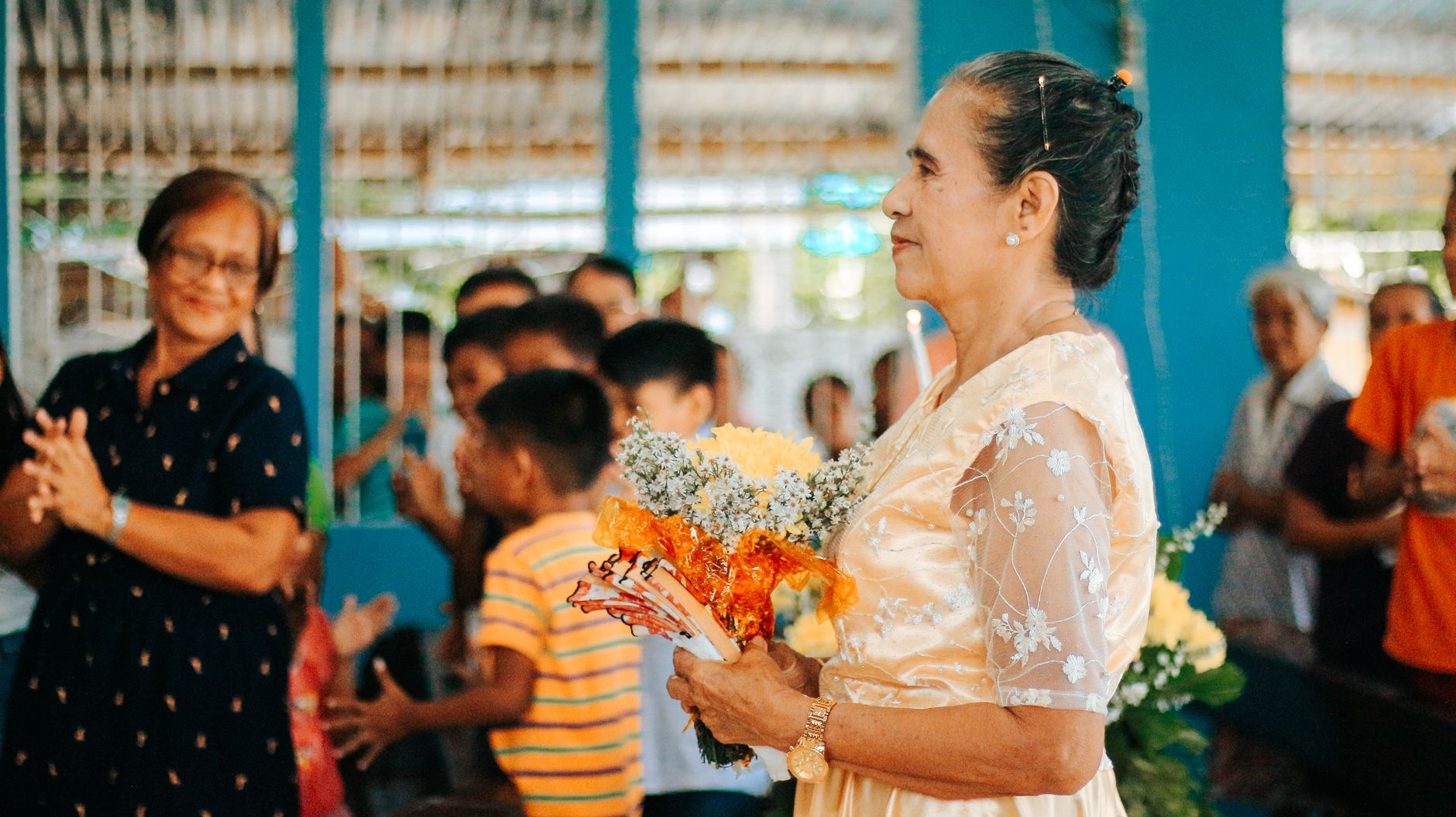 Elderly woman in a light yellow dress holding flowers at an event