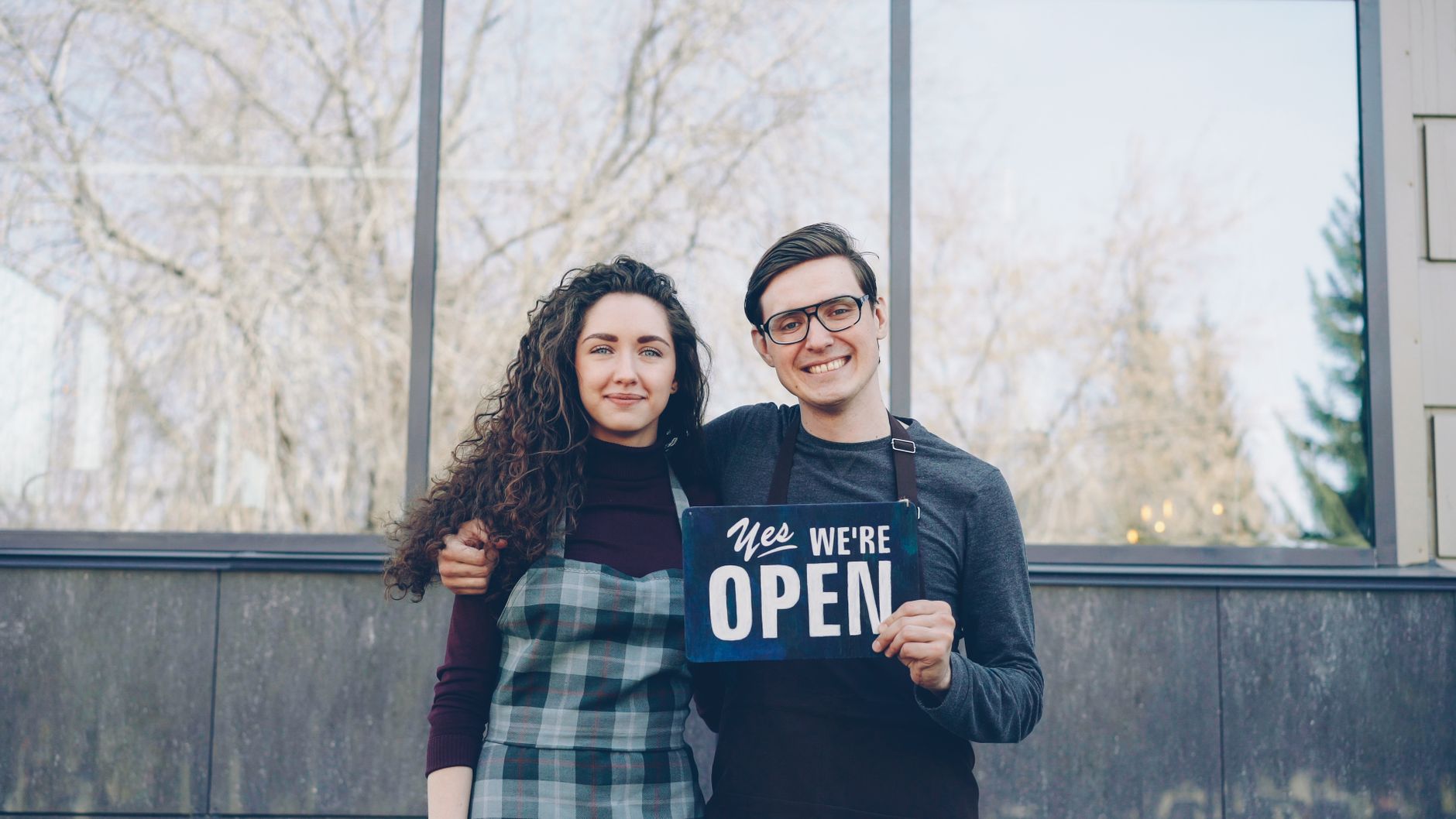 Two smiling business owners hold a "Yes We're Open" sign outside their shop