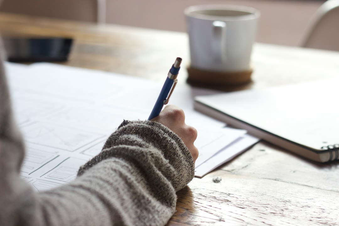 A person's hand in a sweater writes notes on a desk with coffee and notebook