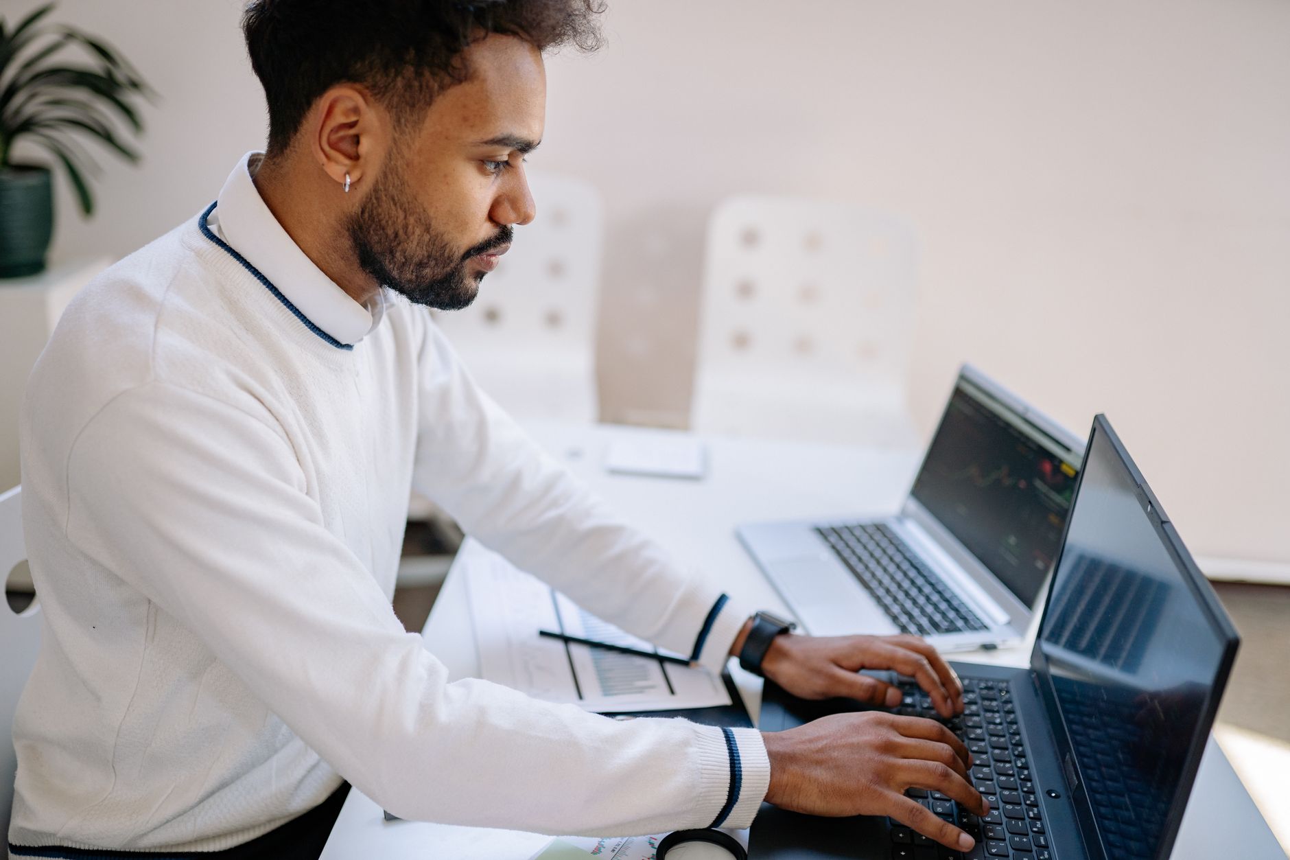 A man sits at a desk typing on a laptop beside another computer
