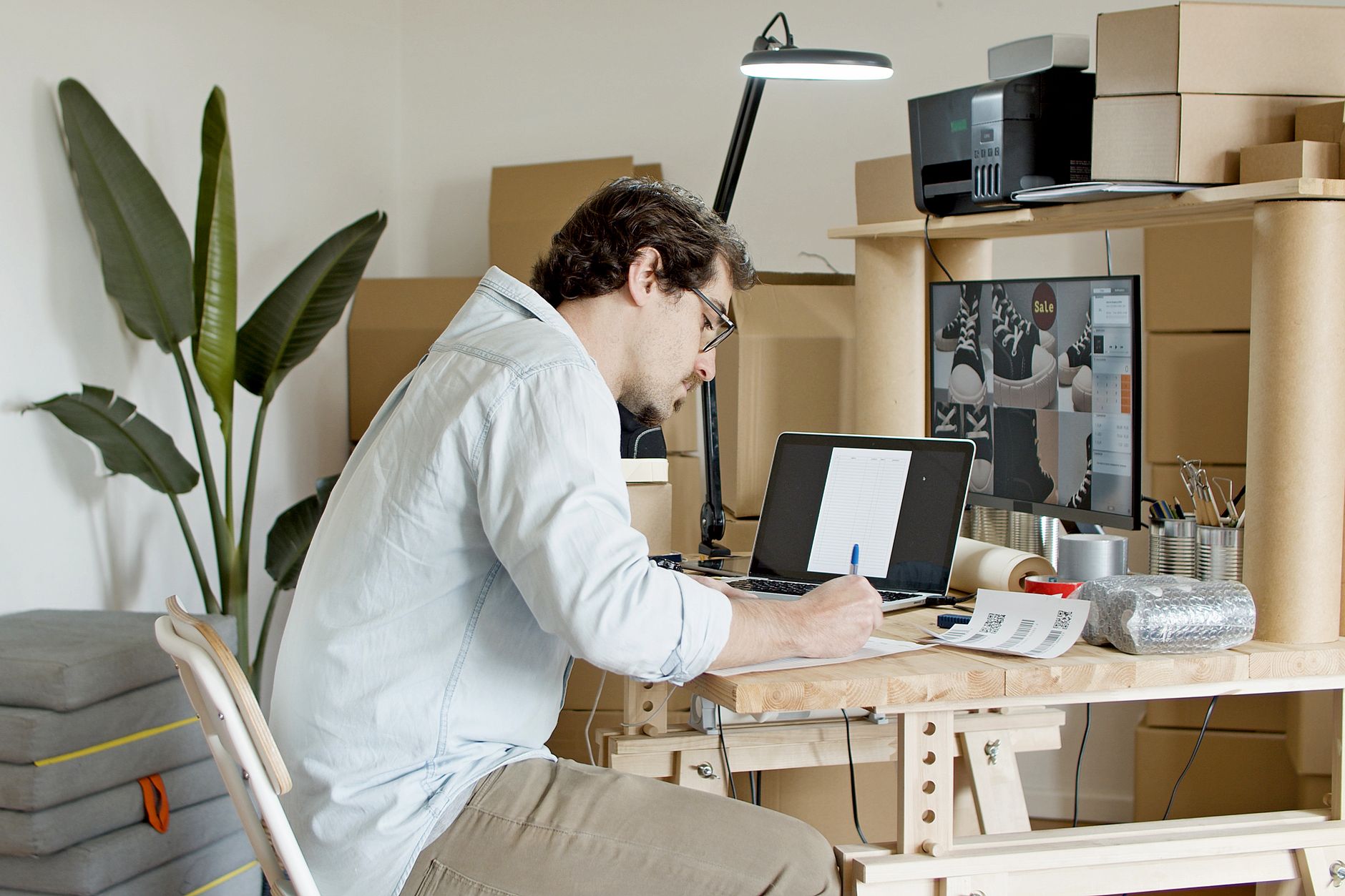 Man working at a desk with laptop, monitor showing shoes, and shipping supplies