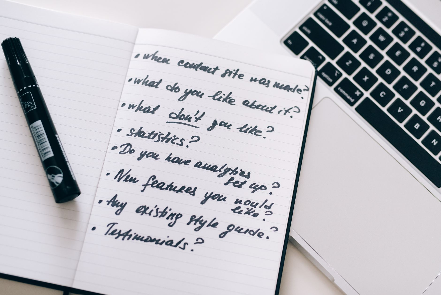 Notebook with handwritten content questions, a pen, and a laptop for scheduling blog posts