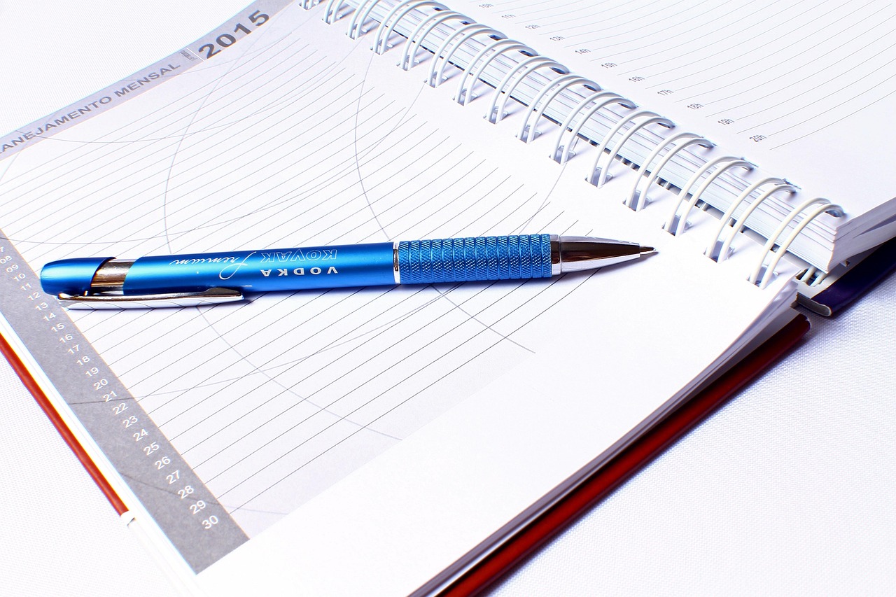A blue pen rests on an open spiral notebook with planning pages for scheduling tasks