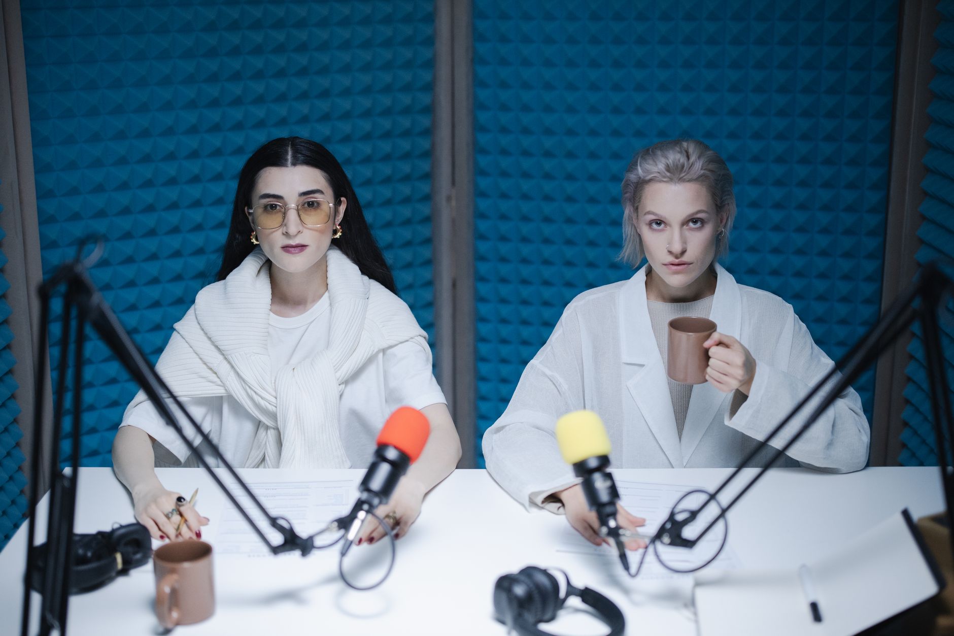 Two women host a podcast in a soundproofed studio with microphones and mugs