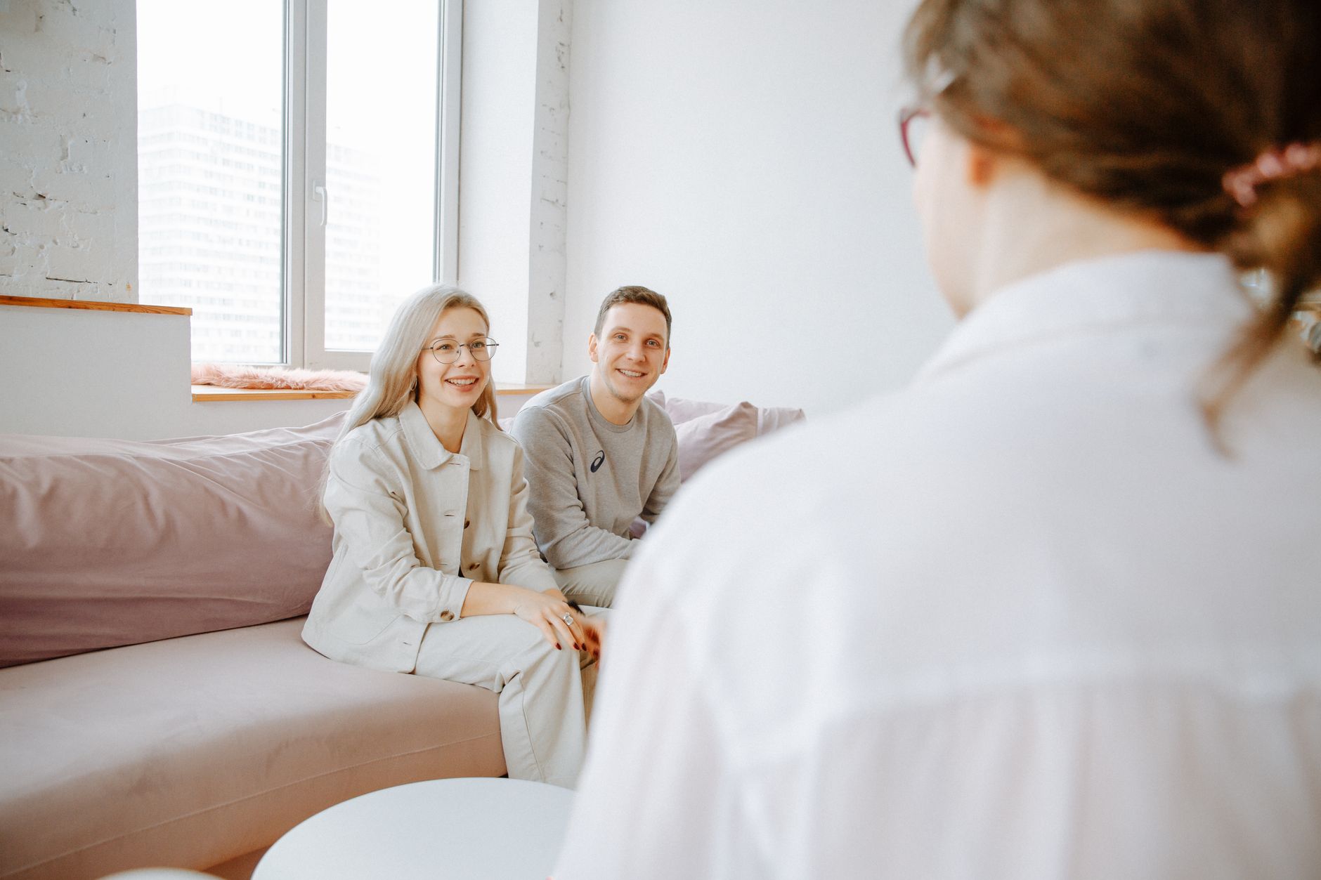 A smiling couple sits on a sofa facing a woman seen from behind