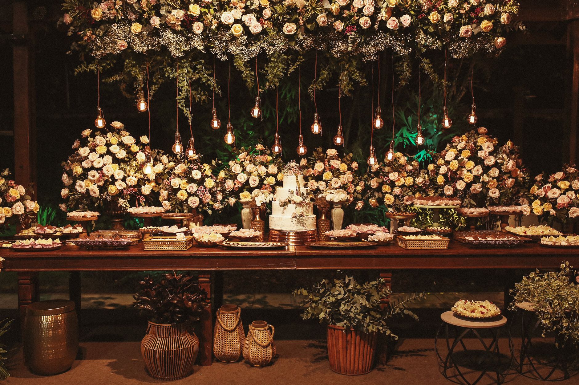 A wedding cake and desserts are on a table decorated with flowers and Edison bulbs