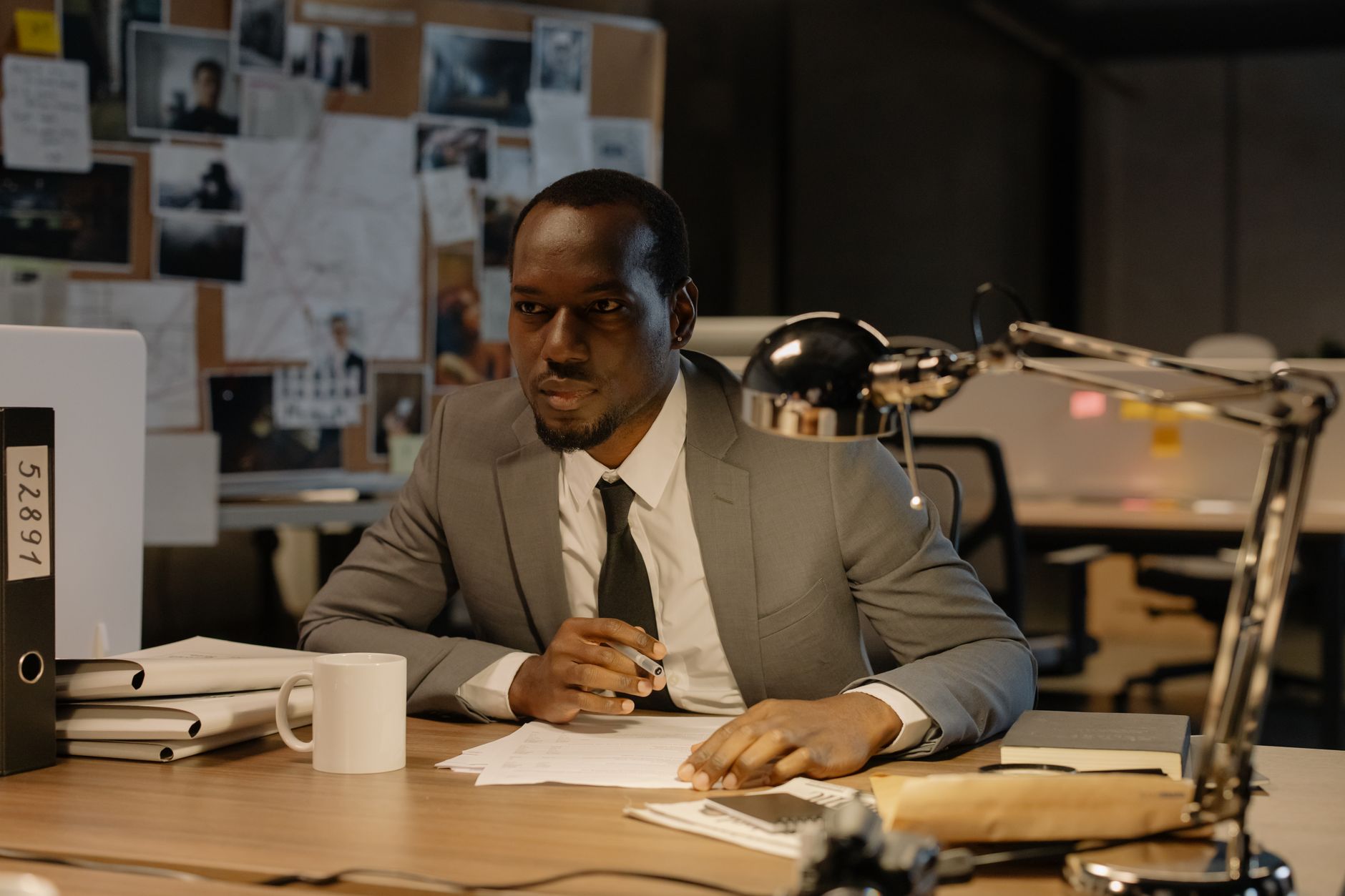 A serious man in a suit works at a desk with case files and documents