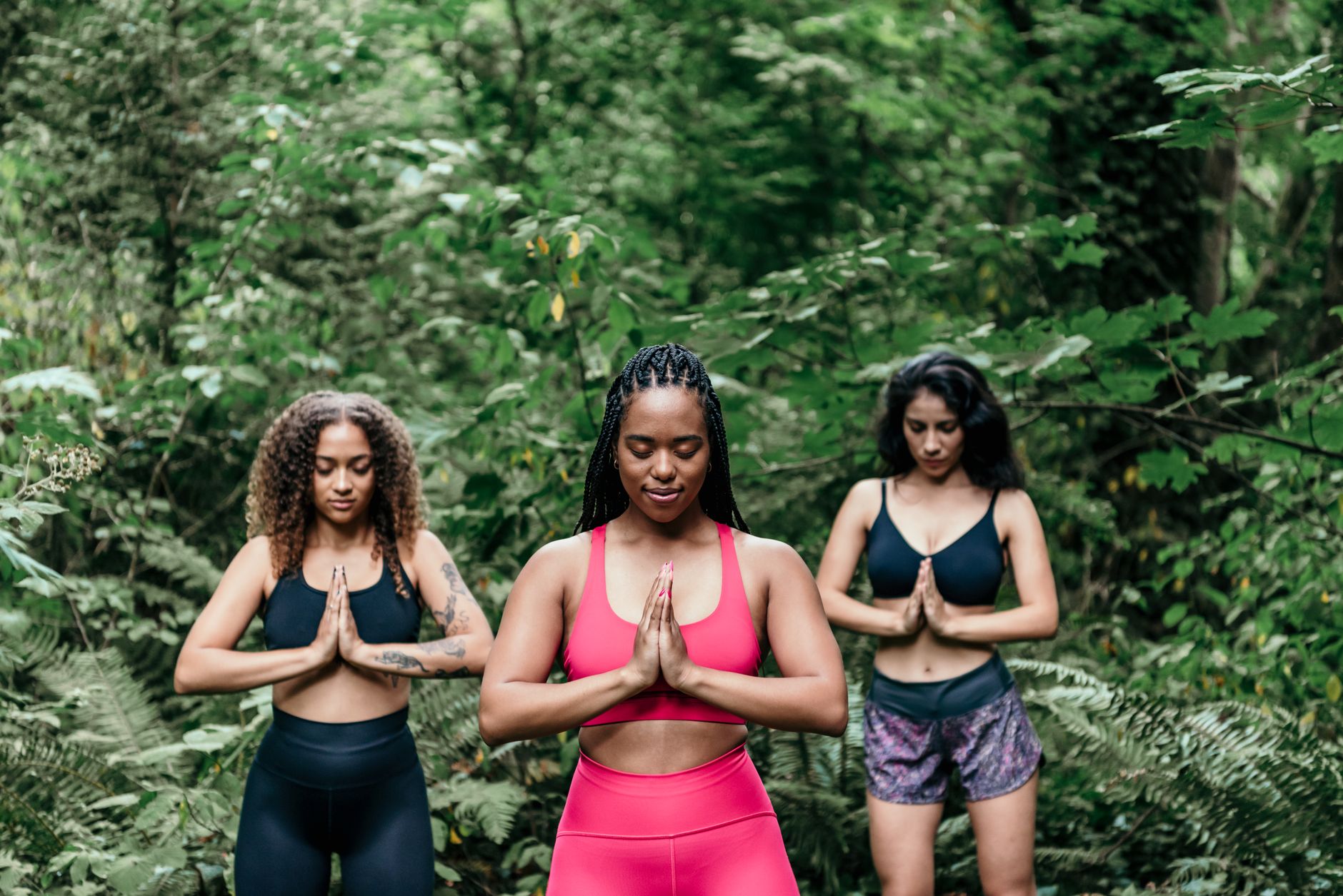 Three women meditate outdoors in a lush green forest, hands in prayer pose