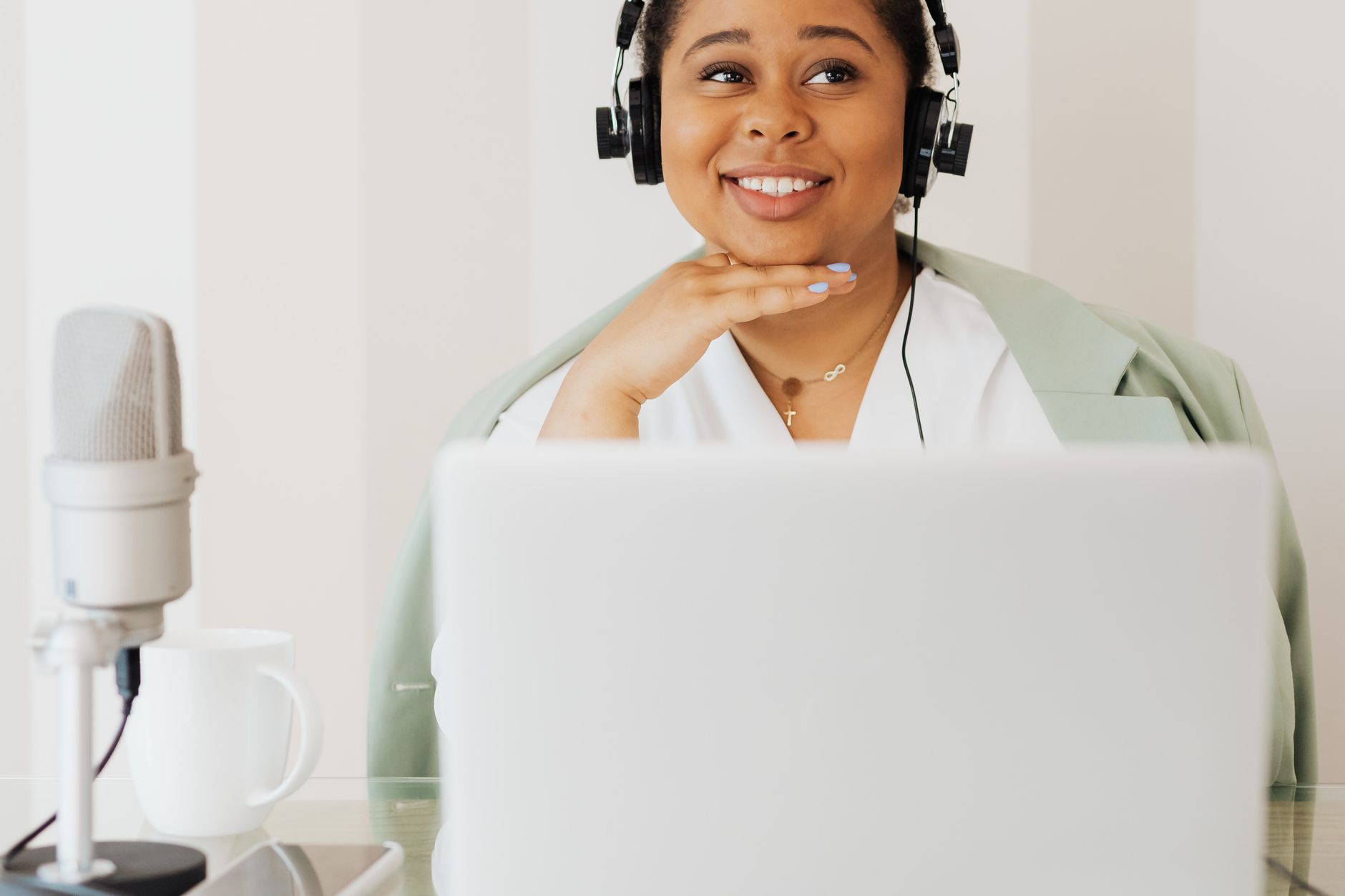 A smiling podcaster in headphones working on a laptop with a microphone nearby