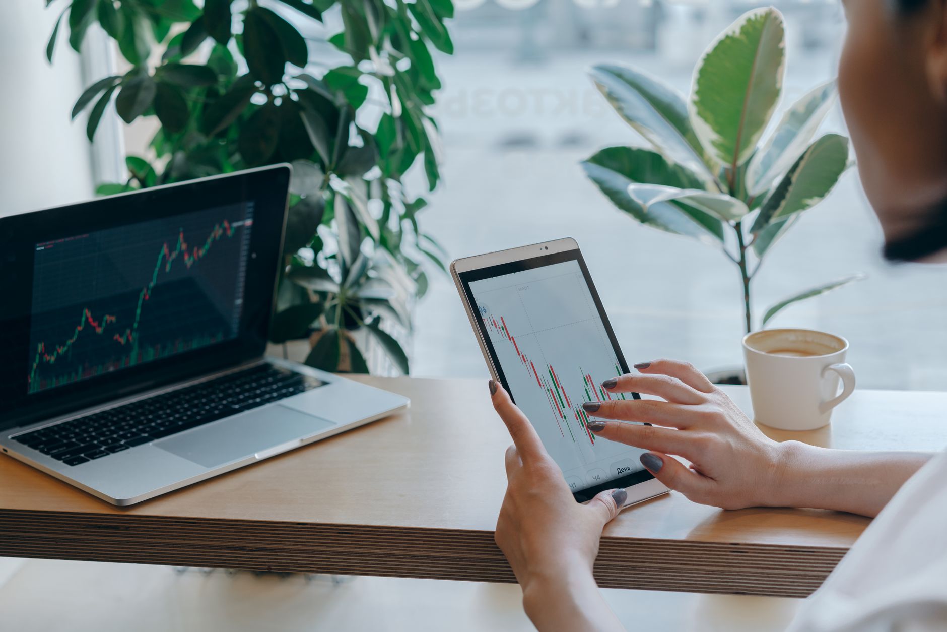 Hands hold a tablet displaying financial charts next to a laptop with similar data