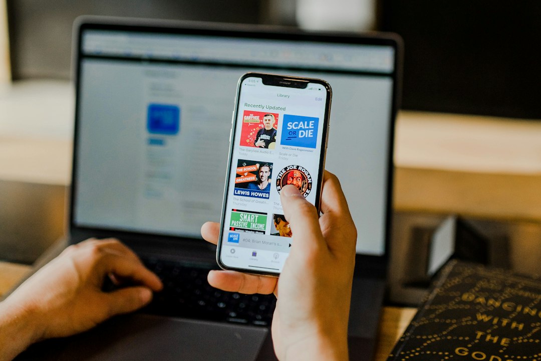 Person browsing podcast episodes on a smartphone in front of an open laptop
