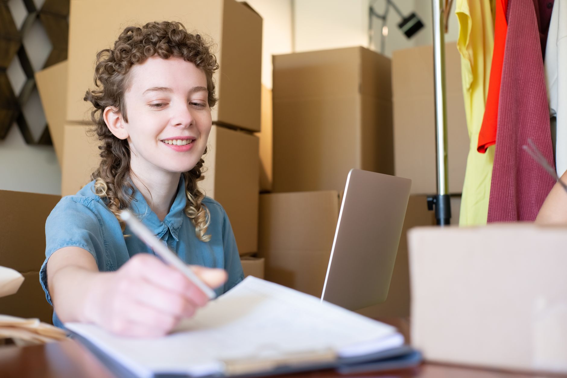 Young woman smiles, writing notes near laptop, surrounded by moving boxes