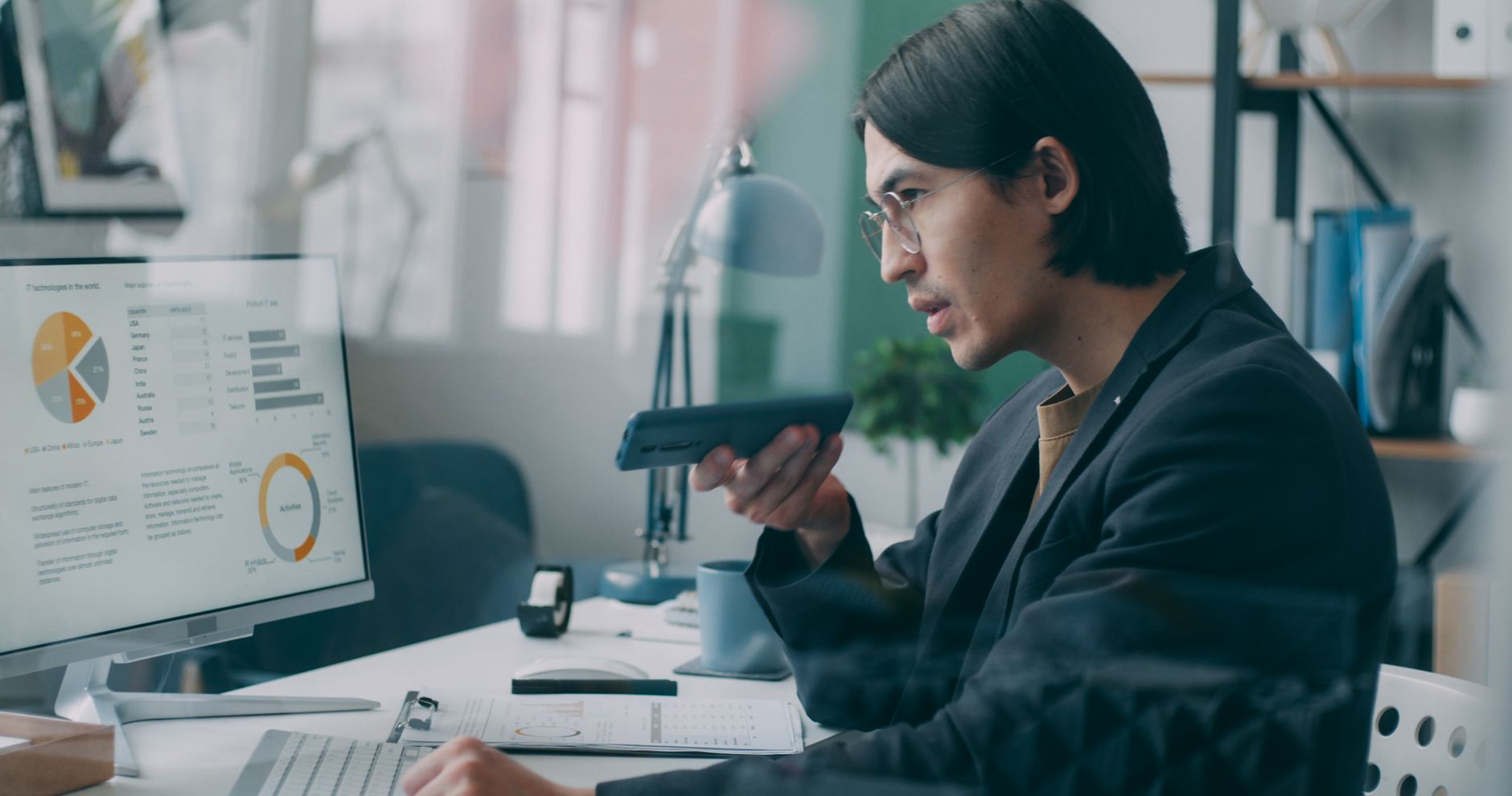 Man looks at data charts on a monitor while holding a smartphone, analyzing podcast growth