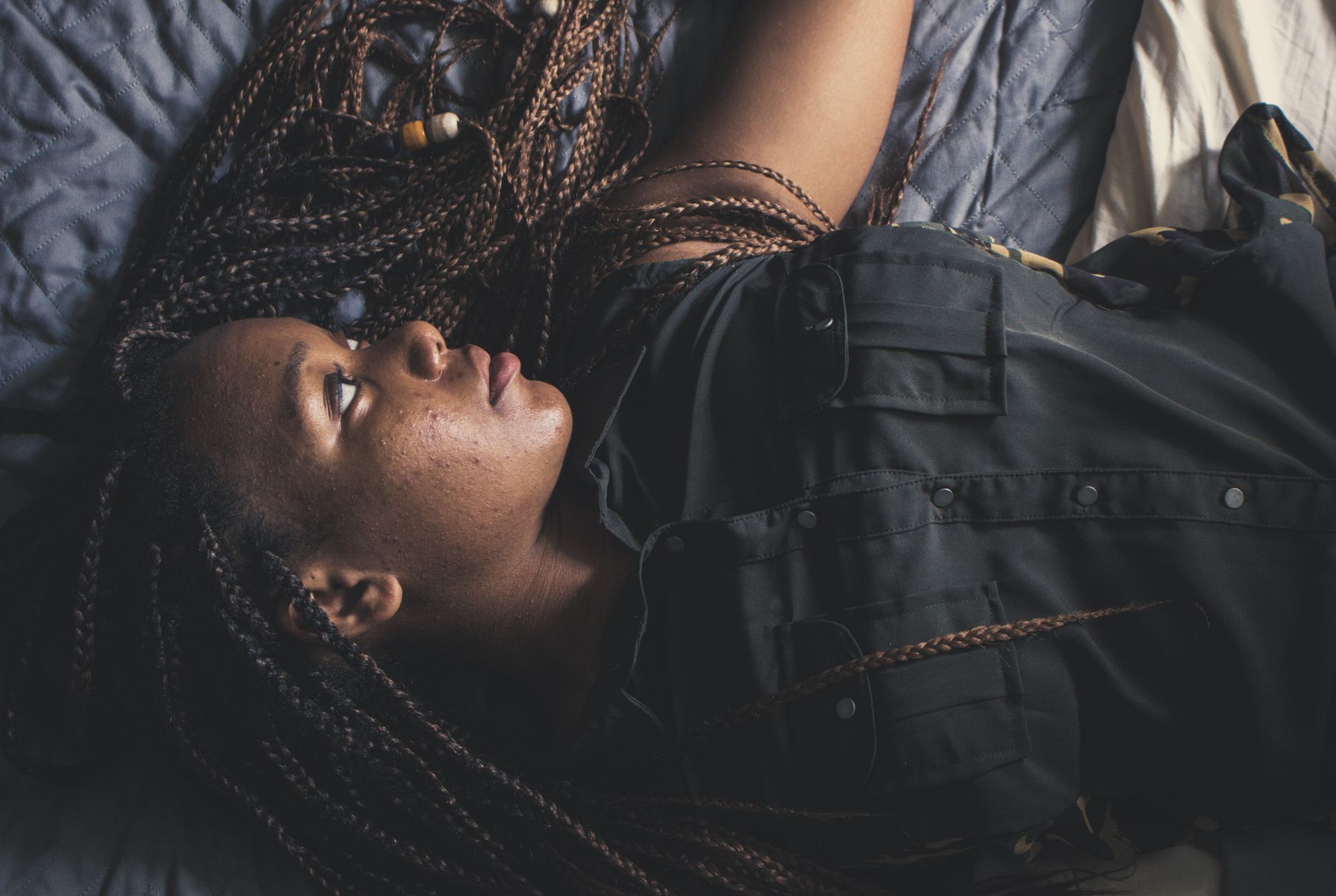A person with braided hair lies on a bed, looking up thoughtfully
