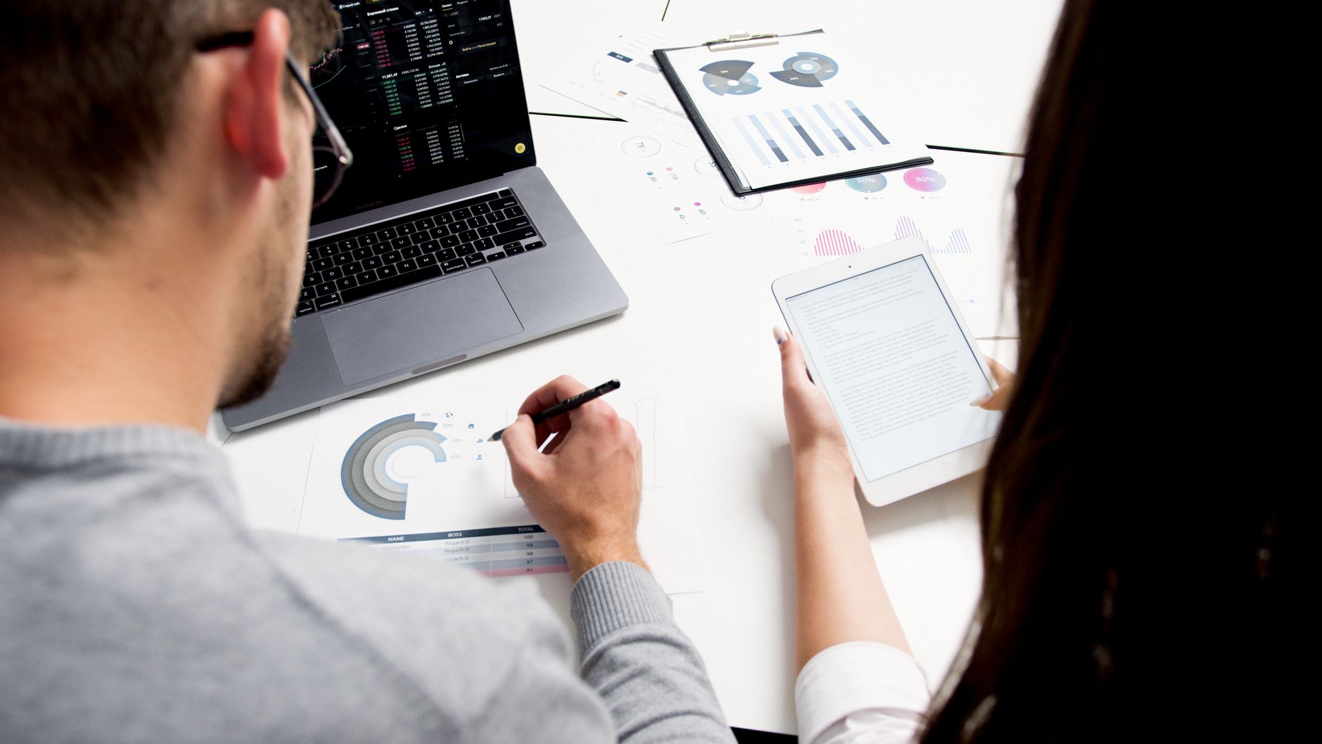 Two people analyzing data on a laptop, tablet, and printed charts